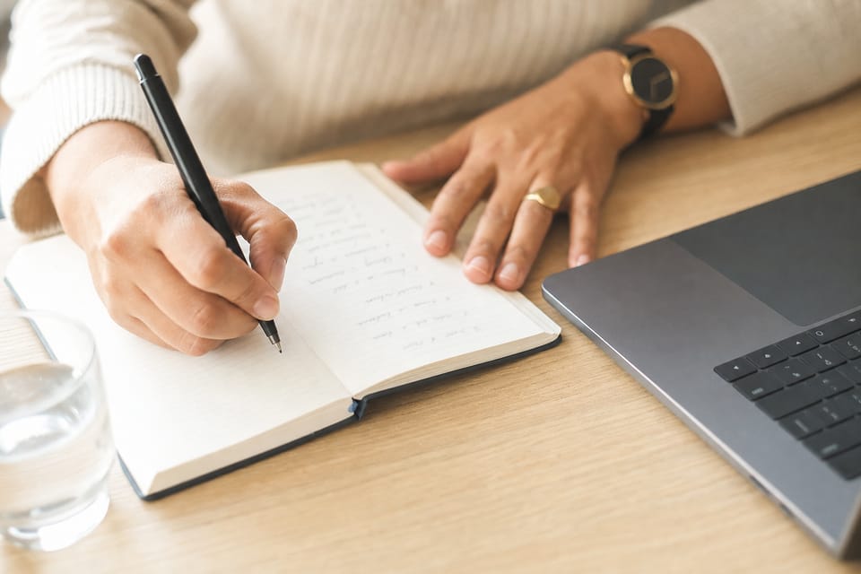 Hands writing a numbered list in a notebook on a pale oak desk.